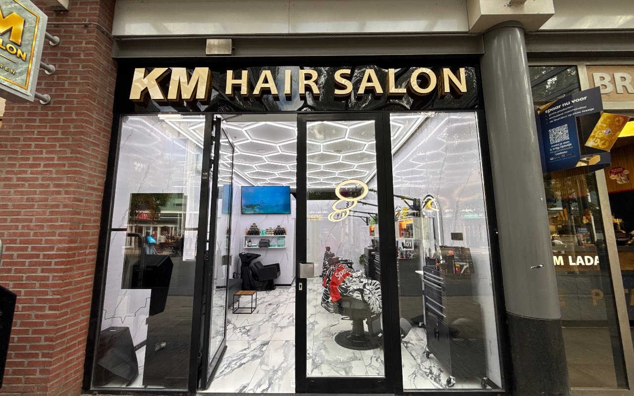 De gevel van KM Hair Salon met gouden letters op een zwart bord en het verlichte zeshoekenplafond achter het glas